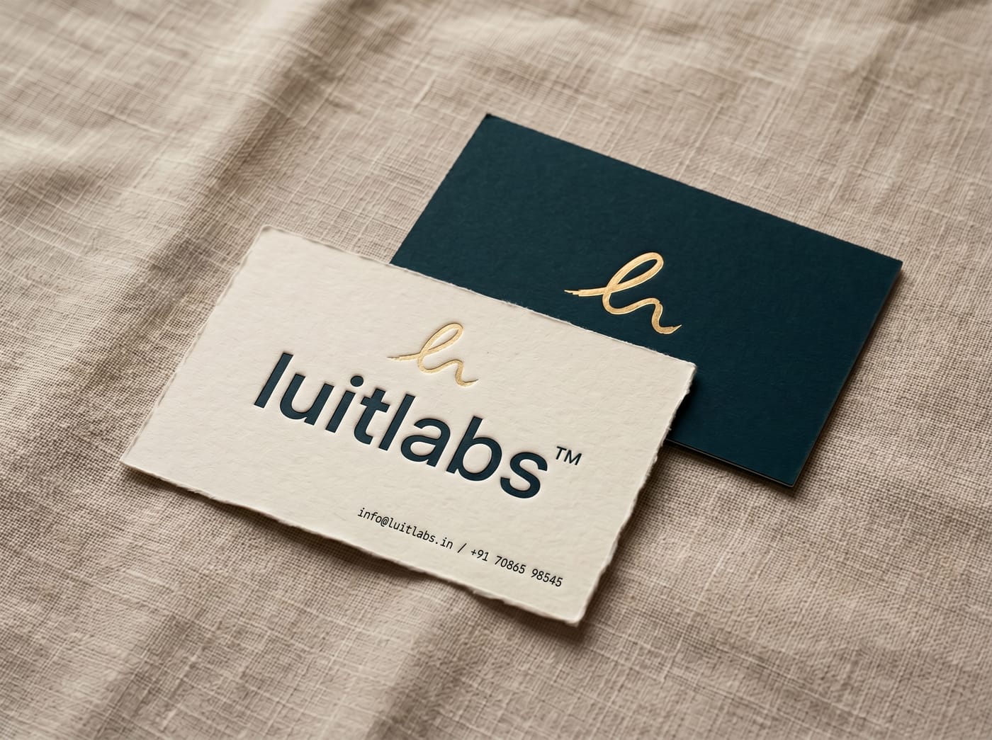 two luitlabs business cards on warm linen — a cream deckle-edge card with the luitlabs wordmark debossed in deep teal and the gold ll mark in foil above; behind it, a dark teal card showing the gold ll mark